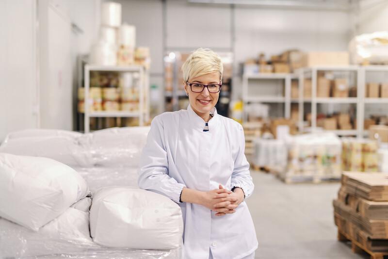 Portrait of smiling young blonde Caucasian blonde woman in sterile uniform and eyeglasses leaning on flour sacks in food factory.