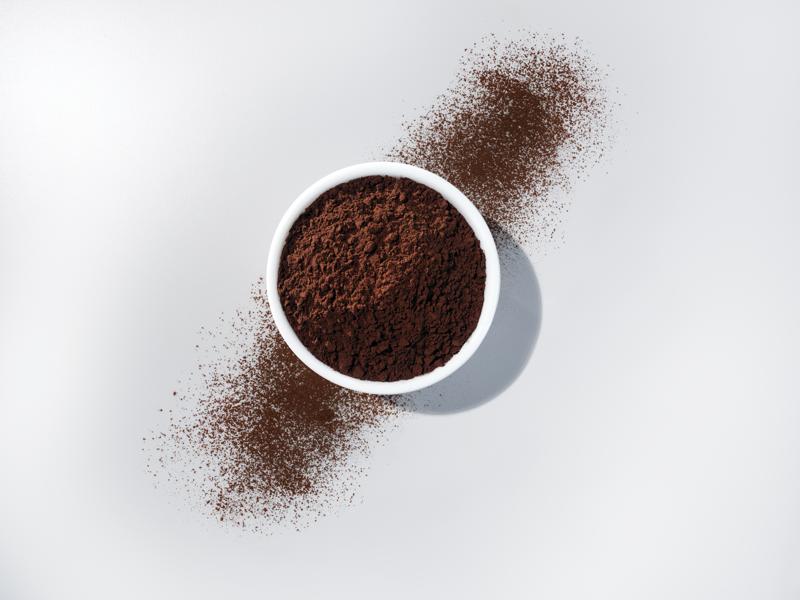 Overhead view of a white bowl filled to the top with a dark brown powder. To either side, powder is scattered diagonally away from the bowl. 