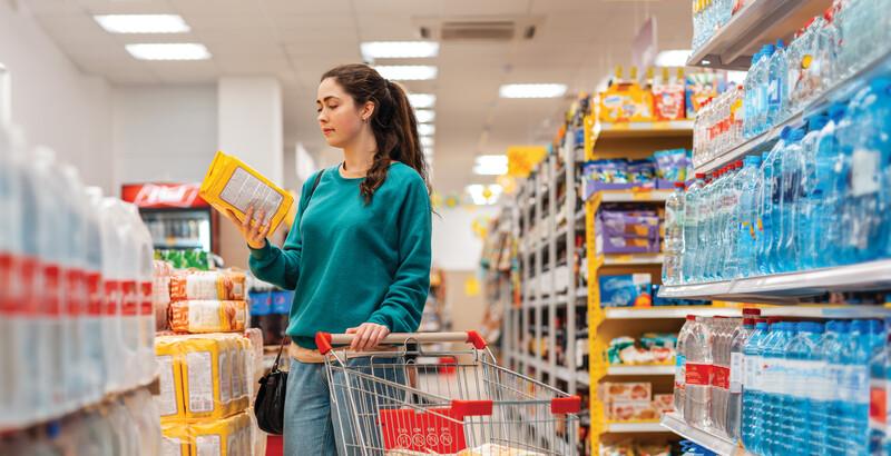 A young pretty Caucasian woman with a grocery cart, reading the ingredients on the package. Store shelves in a blur, in the background. The concept of buying goods and shopping.