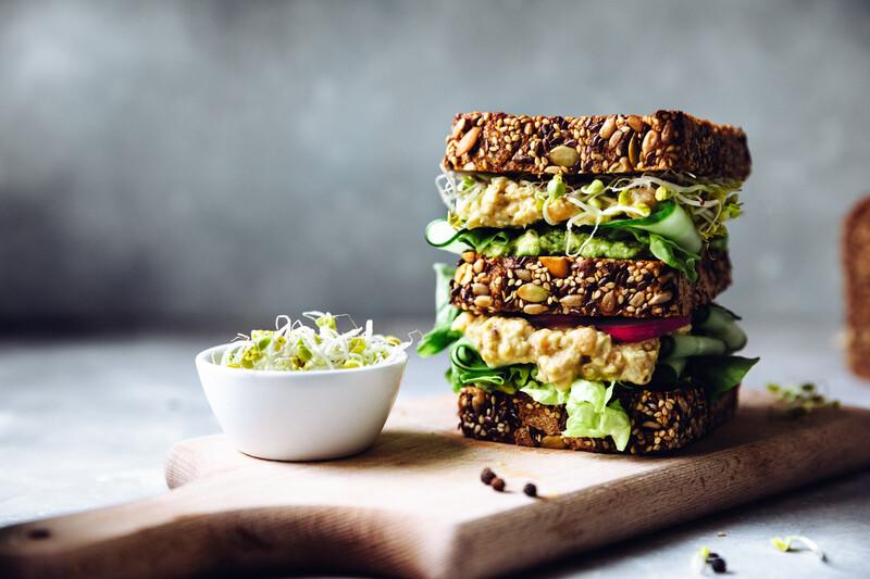 Vegetarian sandwich made with sourdough bread, chickpeas, avocado creme, cucumber, radish and remoulade sauce with bowl of sprouts served on a table.